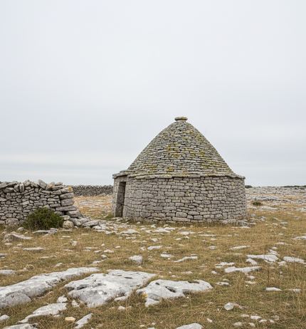 A round drystone hut with a conical lichen mottled roof, a low broken wall to its left, limestone slabs bare in dry grass