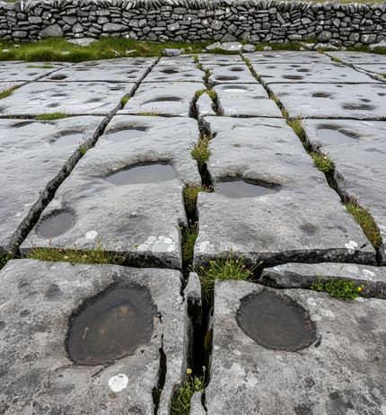 Flat limestone slabs cut by deep grass-filled cracks, dark rainwater pools in worn hollows, a dry stone wall along the far edge