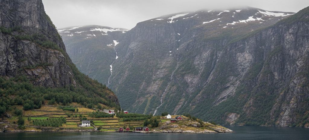 Steep grey fjord walls under low cloud, snow patches high up, thin waterfalls, red and white houses on a green shelf above dark water