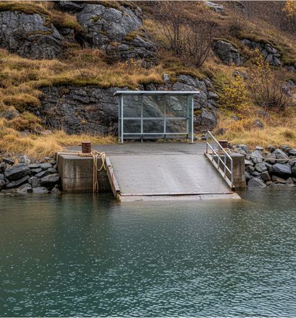 Glass sided waiting shelter beside a concrete ramp sloping into still green grey fjord water, below a rocky slope of tawny autumn grass