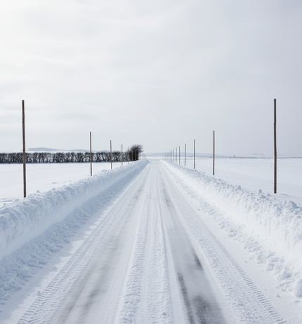 A wide packed snow road running dead straight and level to a flat horizon, tall marker poles along both ploughed verges