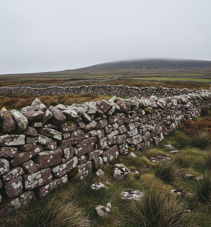 A mortarless drystone wall of reddish blocks crusted with pale lichen, tussocky grass either side, a second wall line and a mist-capped hill beyond