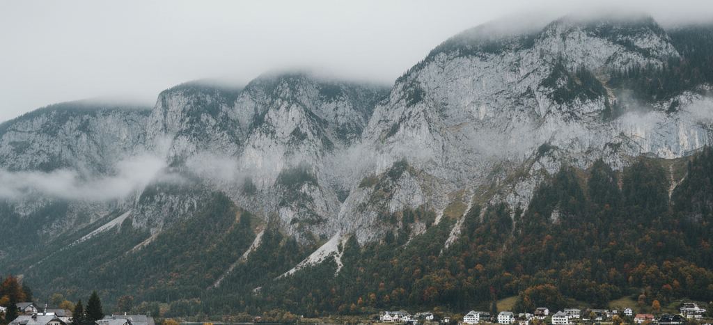 Low cloud along a grey limestone massif, wooded slopes turning rust below it, and a line of white houses at the water